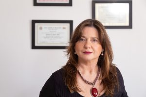 mulher em frente a quadros com certificados usando roupa preta e colar vermelho